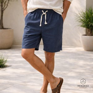 May include: Navy blue shorts with a drawstring waist. The shorts have pockets and a small white tag on the side. The model is wearing a white long-sleeve shirt and brown sandals. The shorts are made of a textured fabric.