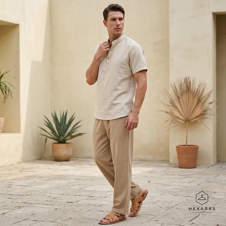 May include: A light beige linen shirt with a mandarin collar and short sleeves, worn with matching beige linen trousers. The outfit is completed with brown leather sandals. The image is taken outdoors.