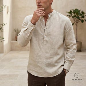 May include: A light beige linen shirt with a mandarin collar and long sleeves. The shirt has a button-down placket and buttoned cuffs. The model is wearing brown pants. The logo for Hexaras Clothing Shop is in the bottom right corner.