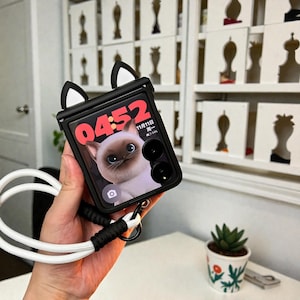 May include: Black phone case with cat ears and a white lanyard. The phone screen displays a cat image and the time 04:52. The case is held in a hand.