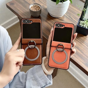 May include: Two leather phone cases with ring holders. One case is brown with a black interior, the other is orange. Both cases have a silver ring holder and a black border. The cases are designed to fold open.