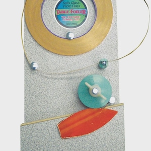 May include: Abstract art piece featuring a gold-colored circular element with the text "TABLE FOILER" and "Foils Glass - First Class". Other elements include a teal disc, an orange leaf shape, and silver spheres.