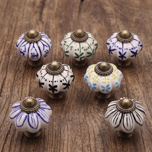 Op de afbeelding: Zeven keramische kastknoppen met een gegroefde, pompoenachtige vorm. Elke knop heeft een witte basis met verschillende handgeschilderde patronen in blauw, groen, zwart of geel, afgewerkt met een centrale dop in antiek messingkleurig metaal.
