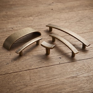May include: A collection of five brass-toned cabinet hardware pieces, including four arched handles and one small T-shaped knob. Each piece features a textured, hammered surface finish and is mounted on two small cylindrical posts for attachment.