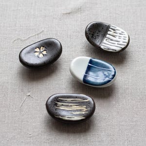 May include: Four oval ceramic chopstick rests with distinct designs. One is black with a gold cherry blossom, one is black with white brushstrokes, one is black and white with textured lines, and one is white with a blue painted center section.