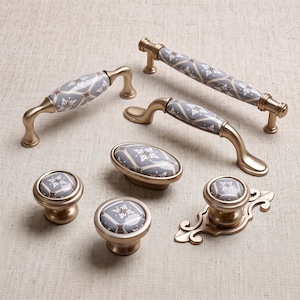 May include: A collection of cabinet hardware including two arched handles, one straight bar handle, two round knobs, one oval knob, and one ornate backplate knob. Each piece features a brushed gold finish and a gray ceramic inlay with a white floral pattern.