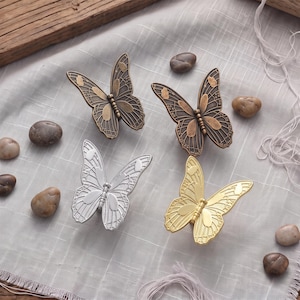 May include: Four butterfly-shaped cabinet knobs in different metallic finishes. One knob is antique bronze, one is dark brown, one is silver, and one is gold. Each knob features detailed wing patterns with raised edges and a central body section.