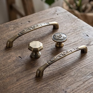 May include: Two arched cabinet pulls and two cabinet knobs in an antique brass finish. The pulls feature a textured surface with an embossed floral vine pattern. One knob is circular with a matching floral design, and the other is octagonal.