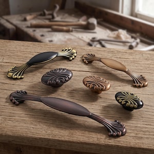 May include: A collection of six metal cabinet hardware pieces including three elongated drawer pulls and three oval knobs. The hardware features a mix of matte black, antique bronze, and copper finishes with ornate, fan-shaped decorative ends.