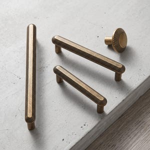 May include: A collection of brass-toned cabinet hardware including three elongated bar pulls and one circular knob. Each piece features a textured, cross-hatched pattern along the sides and a smooth, flat centre strip for a tactile grip.