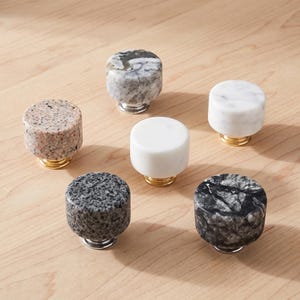 Puede incluir: Una colección de seis pomos cilíndricos para armarios con diversas texturas de piedra, incluyendo beige moteado, mármol gris, blanco sólido, granito gris oscuro y mármol veteado negro, cada uno montado sobre una pequeña base metálica.