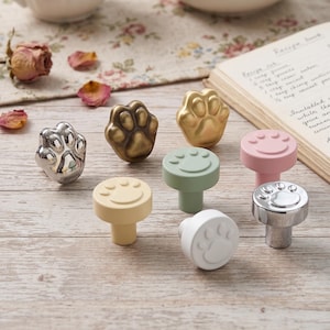 May include: A collection of seven cabinet knobs featuring paw print designs. The set includes metallic silver, antique bronze, and matte gold paw-shaped knobs, alongside round knobs in pastel yellow, sage green, soft pink, and white with embossed paw prints.