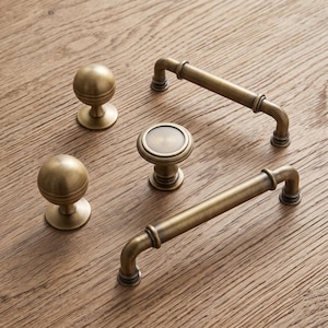 May include: A collection of five brass-toned metal cabinet hardware pieces, including two spherical knobs with horizontal grooves, two tubular bar pulls with rounded ends, and one circular knob featuring a flat, recessed center detail.