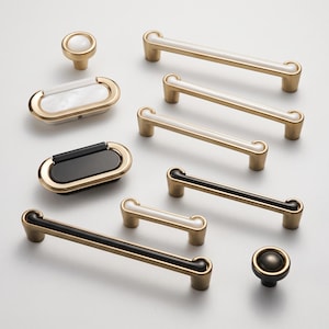 Puede incluir: Una colección de herrajes para muebles que incluye varios tiradores de cajón y pomos. Las piezas presentan un acabado de metal dorado cepillado con inserciones contrastantes en blanco o negro. El conjunto incluye tiradores de barra largos.
