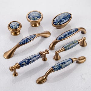 May include: A collection of seven cabinet hardware pieces featuring a brushed brass finish and ceramic inserts. The inserts display a dark blue background with a repeating white and light green wave pattern. The set includes round knobs, oval knobs, and various curved and straight drawer pulls.