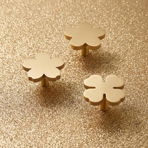 May include: Three gold-toned metal cabinet knobs with a brushed finish. Two of the knobs are shaped like five-petaled flowers, and one knob is shaped like a four-leaf clover. Each knob features a central cylindrical post for mounting purposes.