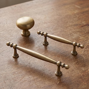 May include: A set of hardware consisting of one round, mushroom-shaped cabinet knob and two elongated, horizontal cabinet pulls. All three pieces are made of a brushed, antique brass-toned metal with a smooth, uniform finish and rounded ends.