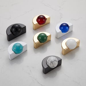 May include: A collection of seven semi-circular cabinet knobs, each featuring a spherical center. The set includes gold, black, and white bases paired with spheres in red, blue, green, turquoise, white, black, and white marble-patterned finishes.