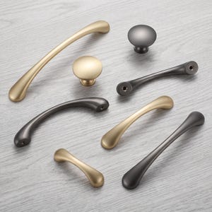 May include: A collection of cabinet hardware including curved drawer pulls and round knobs in matte gold and dark charcoal finishes. The set features various lengths of arched handles and circular knobs, each with visible screw holes for mounting.