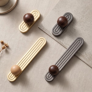 May include: Four cabinet pulls featuring elongated rectangular metal plates with rounded ends and concentric grooved lines. Two pulls are gold-toned with dark wood spheres, one is dark gray with a dark wood sphere, and one is gold with a light wood sphere.