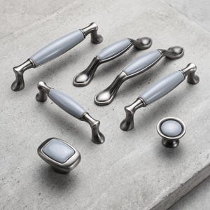Puede incluir: Un conjunto de herrajes para muebles que incluye cuatro tiradores de cajón arqueados, un pomo rectangular y un pomo circular. Cada pieza presenta un acabado metálico oscuro con una inserción de cerámica lisa de color gris claro.