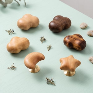 May include: A collection of six wooden cabinet knobs in various shapes including clouds, a crescent moon, and a star. The knobs feature natural wood grain patterns in light and dark brown tones, with two pieces mounted on circular brass bases.