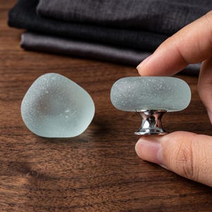 May include: Two cabinet knobs featuring smooth, frosted, pale seafoam green glass tops with a rounded, pebble-like shape. Each knob is attached to a polished, reflective silver-toned metal base that tapers toward the center for mounting.