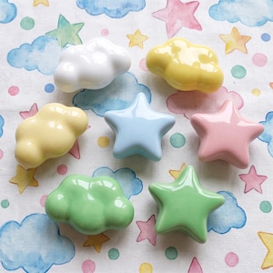 May include: A collection of seven ceramic drawer knobs shaped like clouds and stars in pastel colors including white, yellow, light blue, pink, and light green. Each knob has a smooth, glossy finish and a rounded, three-dimensional form.