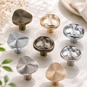 May include: A collection of eight cabinet knobs in various metallic finishes including gold, silver, and bronze. Four knobs feature a concentric spiral pattern, while the other four have a textured, irregular surface with a central indentation.
