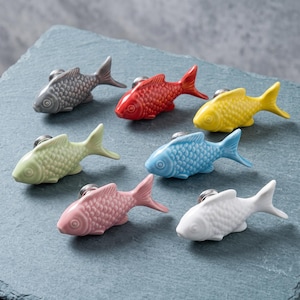 May include: A collection of seven fish-shaped cabinet knobs in various colors including gray, red, yellow, light green, blue, pink, and white. Each knob features a textured scale pattern and a metallic mounting base for attachment to furniture.