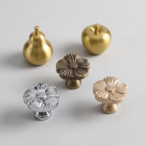 Puede incluir: Un conjunto de cinco pomos para armarios que incluye un pomo dorado con forma de pera, uno con forma de manzana y tres pomos con forma de flor en acabados plateado, latón envejecido y oro rosa. Cada pomo cuenta con un vástago central.