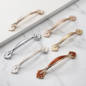 May include: A collection of six curved cabinet handles featuring metallic gold-tone finishes and colored enamel accents in white, soft pink, solid gold, black, and deep orange, each with a decorative fan-shaped detail at both ends.