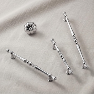 May include: A set of three silver-toned metal cabinet hardware pieces consisting of one round, fluted knob and two elongated drawer pulls. Each pull features a central beaded design with tapered ends and a polished, reflective metallic finish.
