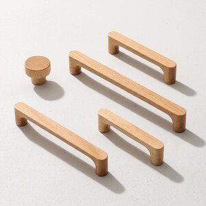 May include: A collection of light-colored wooden cabinet hardware consisting of one circular knob and four rectangular drawer pulls of varying lengths. Each piece features a smooth, natural wood grain finish with rounded edges and a simple design.