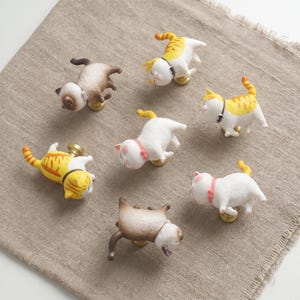 May include: A set of seven cabinet knobs shaped like cats in various poses and colours, including white, orange tabby, and brown Siamese-style patterns. Each cat figure is attached to a small, circular gold-toned metal base for mounting.