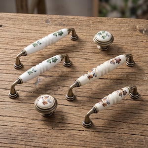May include: A collection of four white ceramic drawer pulls and two round knobs featuring antique brass metal hardware. The pulls have a ribbed centre and are decorated with painted green, blue, and brown leaf patterns on the ceramic surfaces.