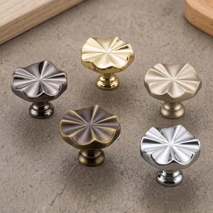 Puede incluir: Cinco pomos de metal para armarios dispuestos en un patrón circular. Cada pomo presenta una forma de flor con crestas radiantes y un tallo central. Los acabados incluyen oro pulido, latón cepillado, bronce oscuro, bronce antiguo y cromo.
