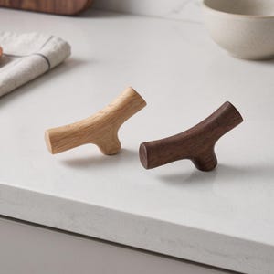 May include: Two wooden cabinet knobs shaped like small tree branches. One knob is crafted from light-colored wood with a visible grain, while the other is made from a darker, rich brown wood. Both feature a smooth, polished finish and a Y-shape.
