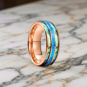 Puede incluir: Anillo de oro rosa con una banda de material iridiscente azul y verde. El anillo tiene un acabado pulido y está sobre una superficie de mármol. El diseño del anillo presenta una franja azul central flanqueada por secciones iridiscentes verdes y azules.