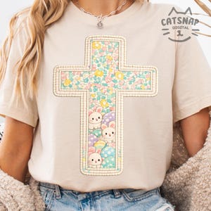 以下が含まれることがあります： クリーム色のTシャツ。花柄とイースターバニーと装飾された卵で満たされた十字のデザインが特徴です。十字はステッチされたボーダーで縁取られています。シャツはクルーネックと半袖です。