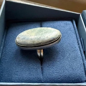 May include: A silver ring featuring a large, oval-shaped, smooth, pale green stone with subtle grey marbling. The stone is set in a simple metal bezel, and the ring band is visible beneath the setting, resting in a dark blue velvet box.