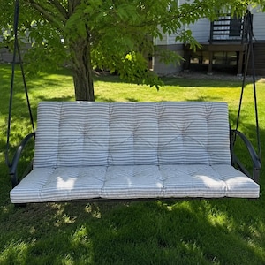 May include: Outdoor swing with a black metal frame and a gray and white striped cushion. The swing is suspended from black chains and is set in a grassy yard with a tree in the background.