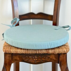 May include: A round, light blue seat cushion with fabric ties on both sides, placed on a wooden chair with a woven seat. The cushion features a smooth, solid-colored top surface and a piped edge, designed to be secured to the chair frame.