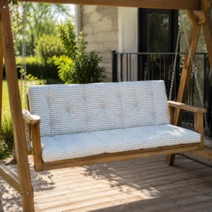 May include: A wooden porch swing with a light gray and white striped cushion. The swing is suspended by chains and has a natural wood finish. The swing is on a wooden deck, with greenery in the background.