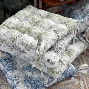 May include: A stack of square seat cushions featuring a white base with green or blue toile de jouy patterns depicting pastoral scenes. Each tufted cushion includes fabric ties at the corners for securing to furniture, with visible stitching.
