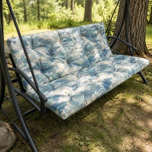 May include: A two-person outdoor swing with a black metal frame. The seat and back cushions feature a blue, green, and white watercolor pattern. The swing is set in a grassy area with trees in the background.