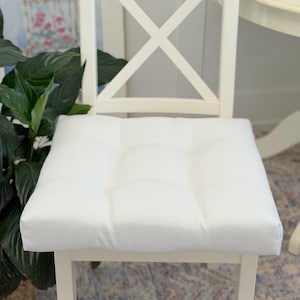 May include: A square, white tufted chair cushion with a textured fabric surface, positioned on a white wooden chair featuring an X-shaped backrest. The cushion has a clean, solid white appearance with visible stitching details at the tufted points.