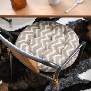 May include: A round, tufted seat cushion featuring a beige and white chevron pattern. The cushion is secured to a metal chair frame with brown fabric ties. The design includes multiple tufted indentations across the circular surface.