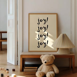 May include: A framed print with the word "joy!" repeated four times in black script. A beige teddy bear sits on the floor near a wooden bench with a stack of books and a lamp. The room has a warm, inviting atmosphere.