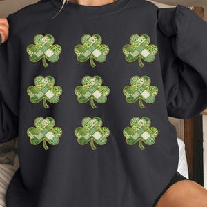 Op de afbeelding: Zwart sweatshirt met negen groene klaverontwerpen. Elke klaver heeft een patchworkpatroon met verschillende tinten groen en wit. De klavers zijn goudkleurig omlijnd, passend bij St. Patrick's Day.